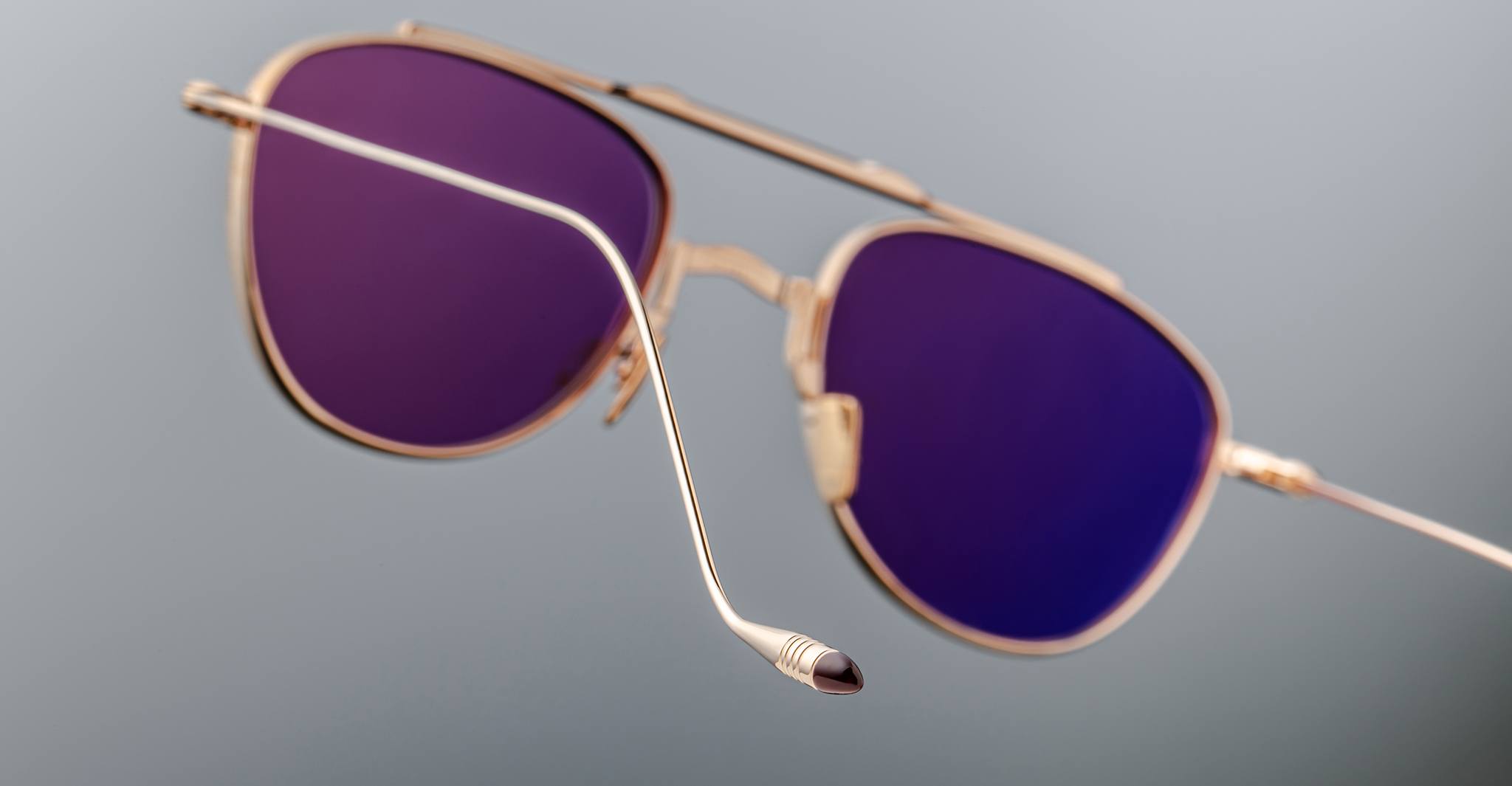 A close-up of Jacques Marie Mage Flynn gold-framed aviator sunglasses with purple-tinted lenses set against a plain gray background, showcasing the detailed design of one prominently featured arm in the foreground.