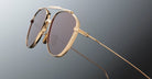Close-up of Jacques Marie Mage Flynn gold metal sunglasses with brown tinted lenses against a gradient gray background. Thin temples and nose pads are visible, emphasizing the eyewears refined design and premium materials.