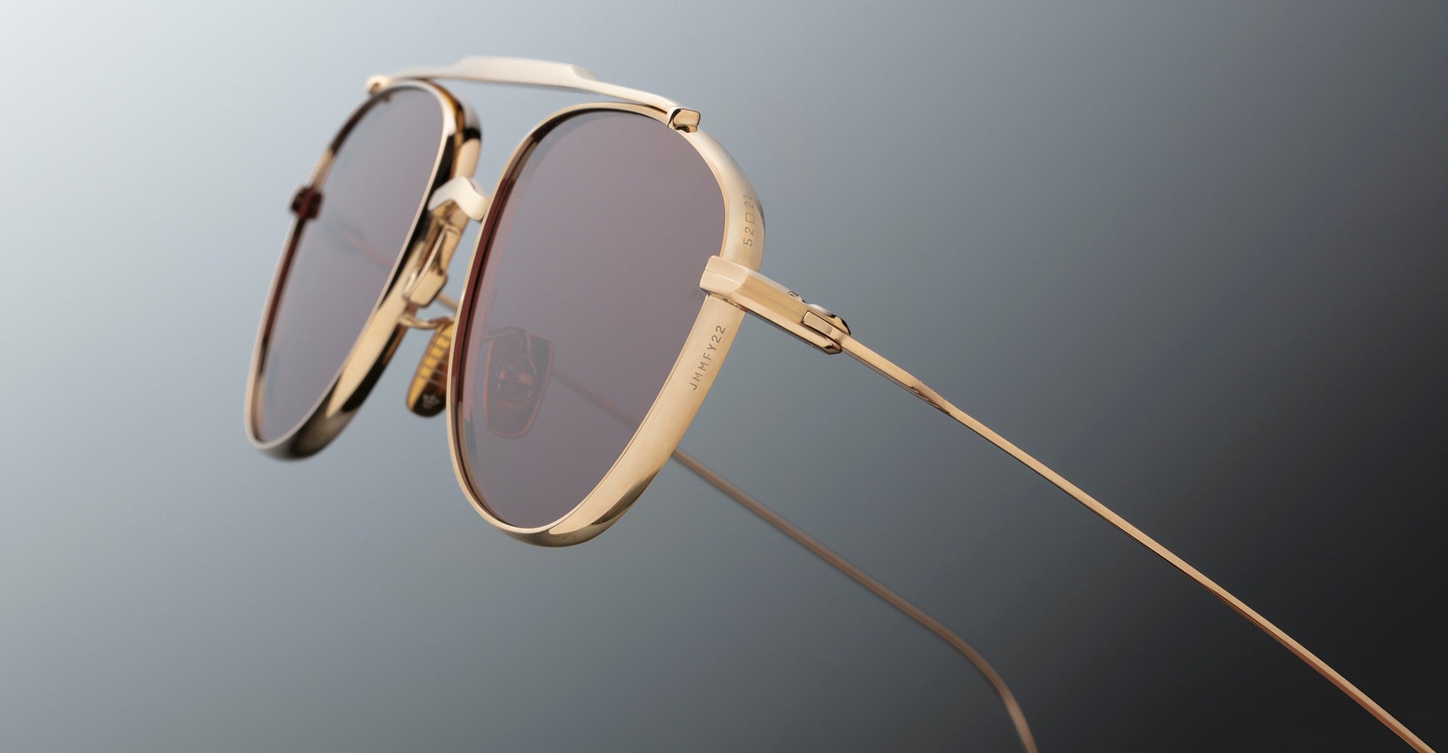 Close-up of Jacques Marie Mage Flynn gold metal sunglasses with brown tinted lenses against a gradient gray background. Thin temples and nose pads are visible, emphasizing the eyewears refined design and premium materials.