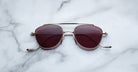 A pair of Jacques Marie Mage Flynn sunglasses with gold frames and dark red lenses rests on a white marble surface.