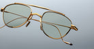 The Jacques Marie Mage Flynn eyeglasses feature gold aviator-style frames and light green tinted lenses, displayed against a plain light gray background.