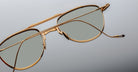 Close-up of Jacques Marie Mage Flynn aviator sunglasses with gold frames and green lenses, placed on a grey surface—one arm extended, the other resting across the lenses.