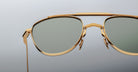 Close-up of the top portion of Jacques Marie Mage Flynn sunglasses, featuring gold frames and green-tinted lenses, set against a plain light gray background.