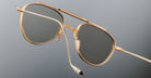A close-up of Jacques Marie Mage Flynn gold-framed aviator sunglasses with dark lenses, suspended against a plain gray background, highlights the detailed curved temple tip.