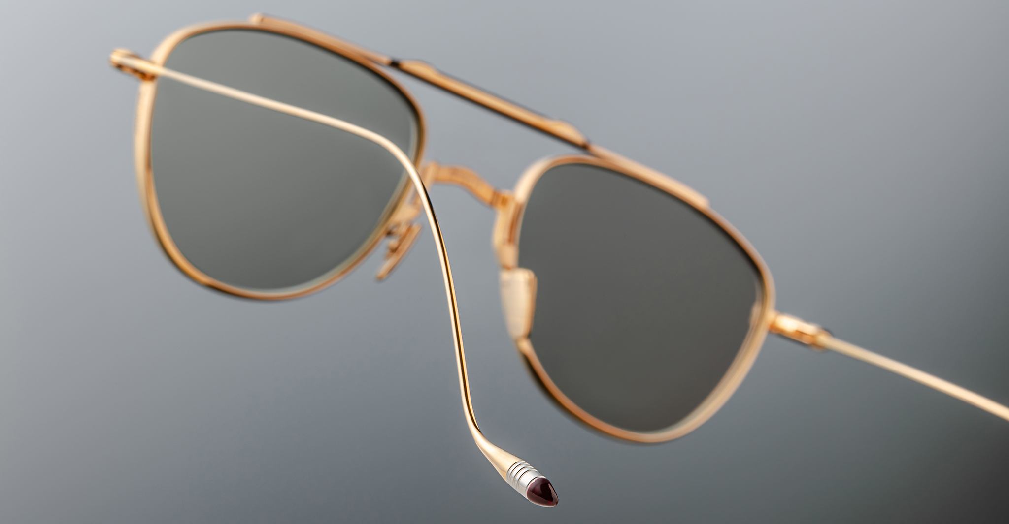 A close-up of Jacques Marie Mage Flynn gold-framed aviator sunglasses with dark lenses, suspended against a plain gray background, highlights the detailed curved temple tip.