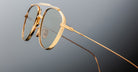 Close-up of Jacques Marie Mage Flynn gold-framed aviator eyeglasses with clear lenses, highlighting detailed design and thin temples against a neutral gray background.