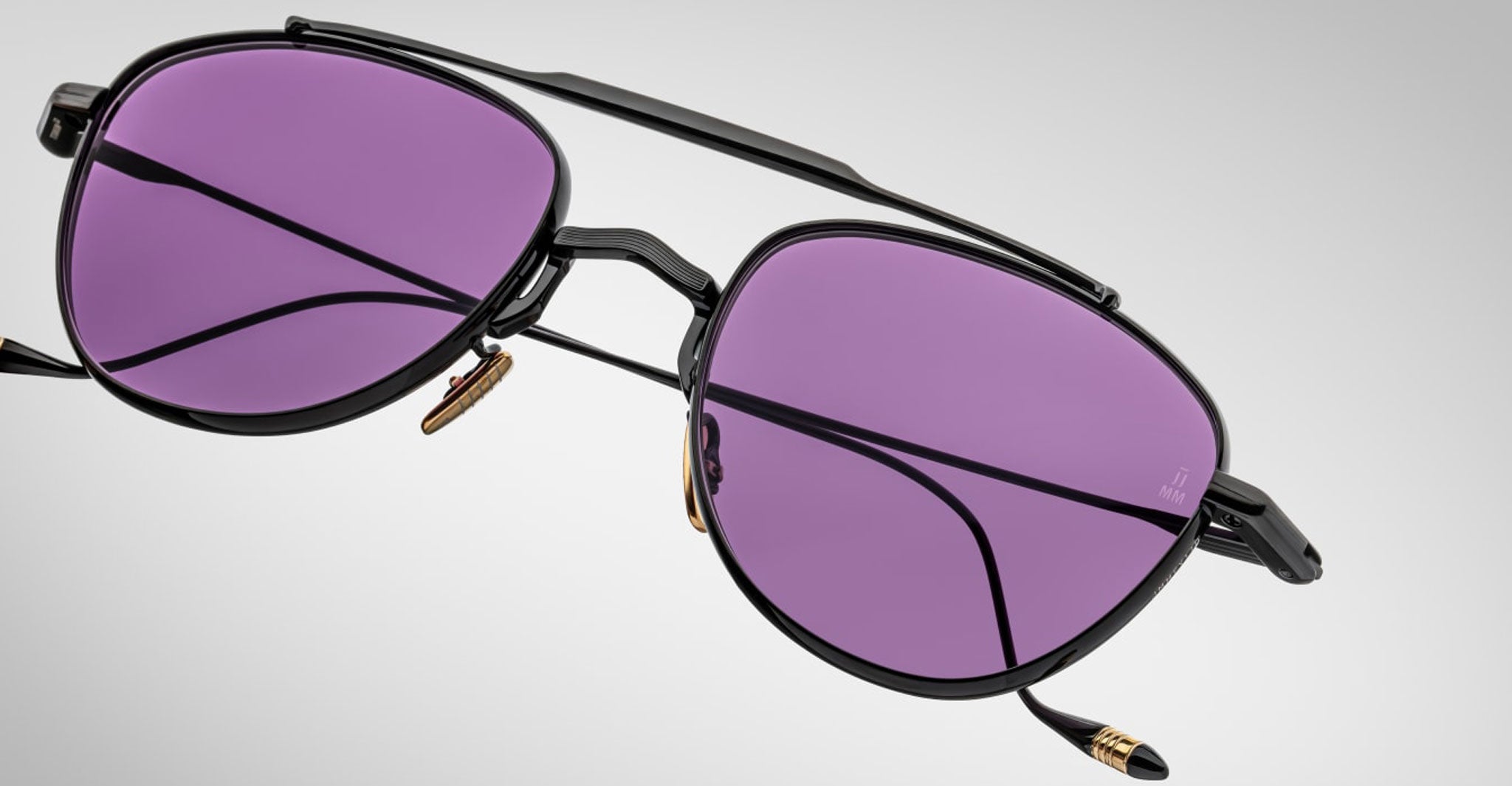 The Jacques Marie Mage Flynn sunglasses feature a black metal aviator-style frame with purple-tinted lenses and gold accents on the nose pads and temple tips, displayed against a plain light gray background.
