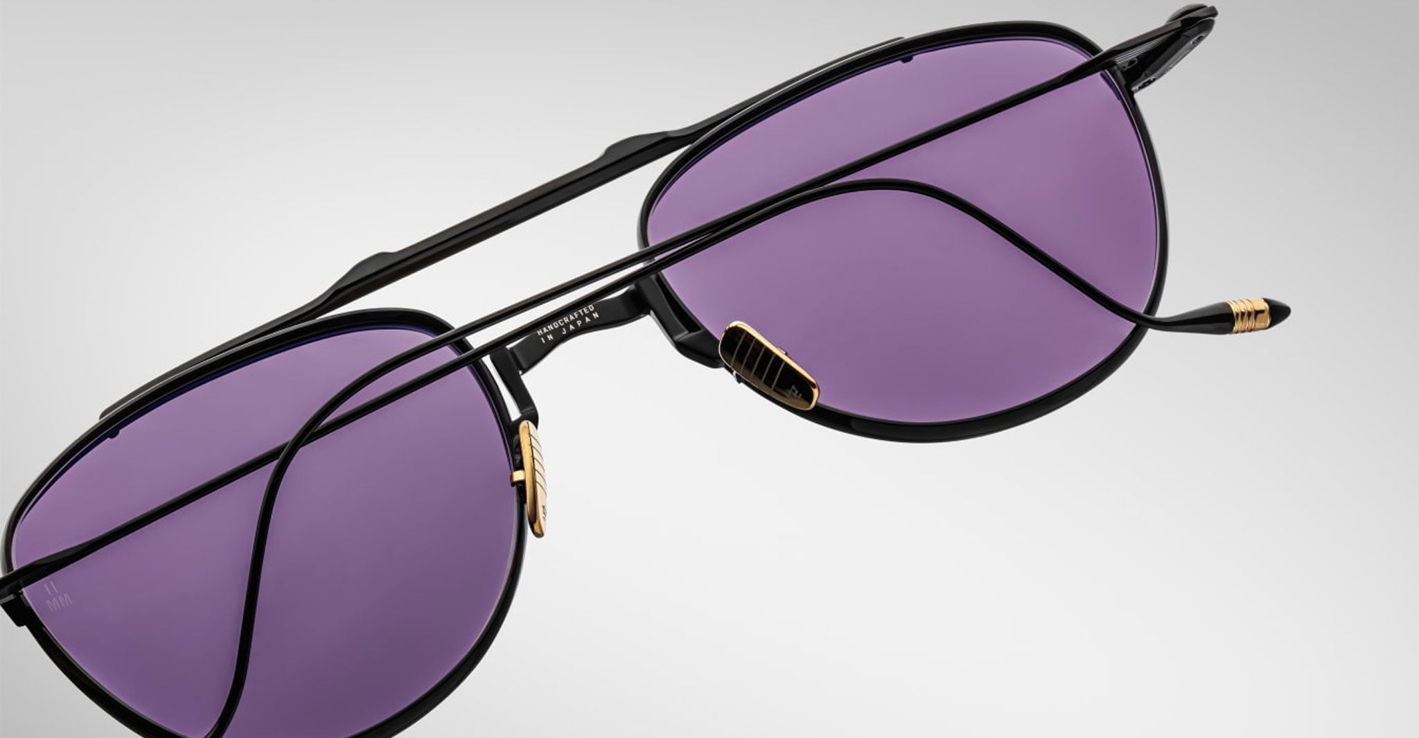 A close-up of Jacques Marie Mage Flynn sunglasses featuring black frames, purple-tinted lenses, and gold accents on the nose pads and temple tips, set against a plain light gray background.