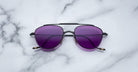 A pair of Jacques Marie Mage Flynn sunglasses with black metal frames and purple-tinted lenses rests on a white marble surface.