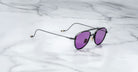 A pair of Jacques Marie Mage Flynn black metal-framed aviator sunglasses with purple-tinted lenses rests on a white marble surface.