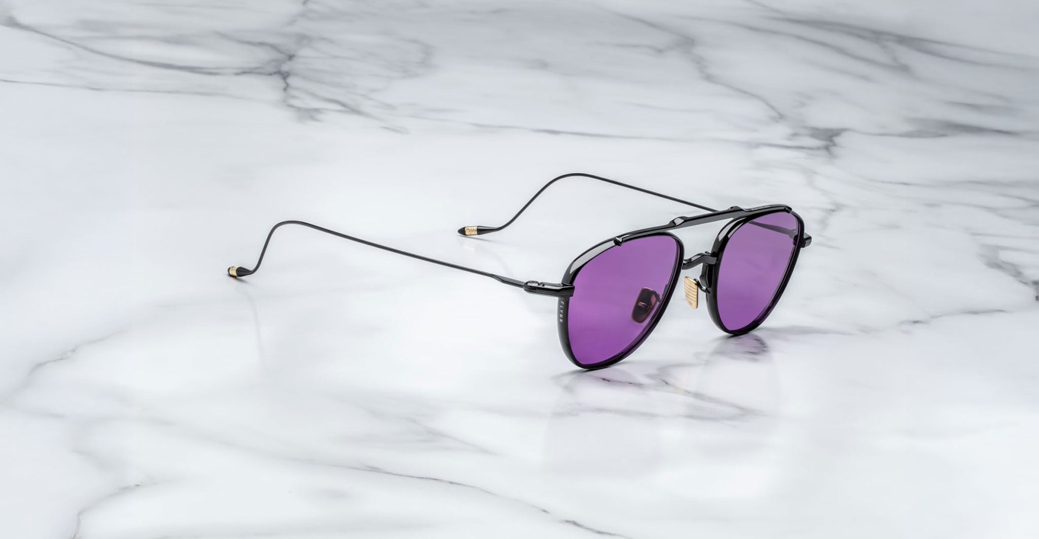 A pair of Jacques Marie Mage Flynn black metal-framed aviator sunglasses with purple-tinted lenses rests on a white marble surface.