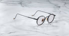 The Jacques Marie Mage Full Metal Jacket eyeglasses, featuring round black rims, thin arms, and gold accents, rest on a white marble surface with gray veining.