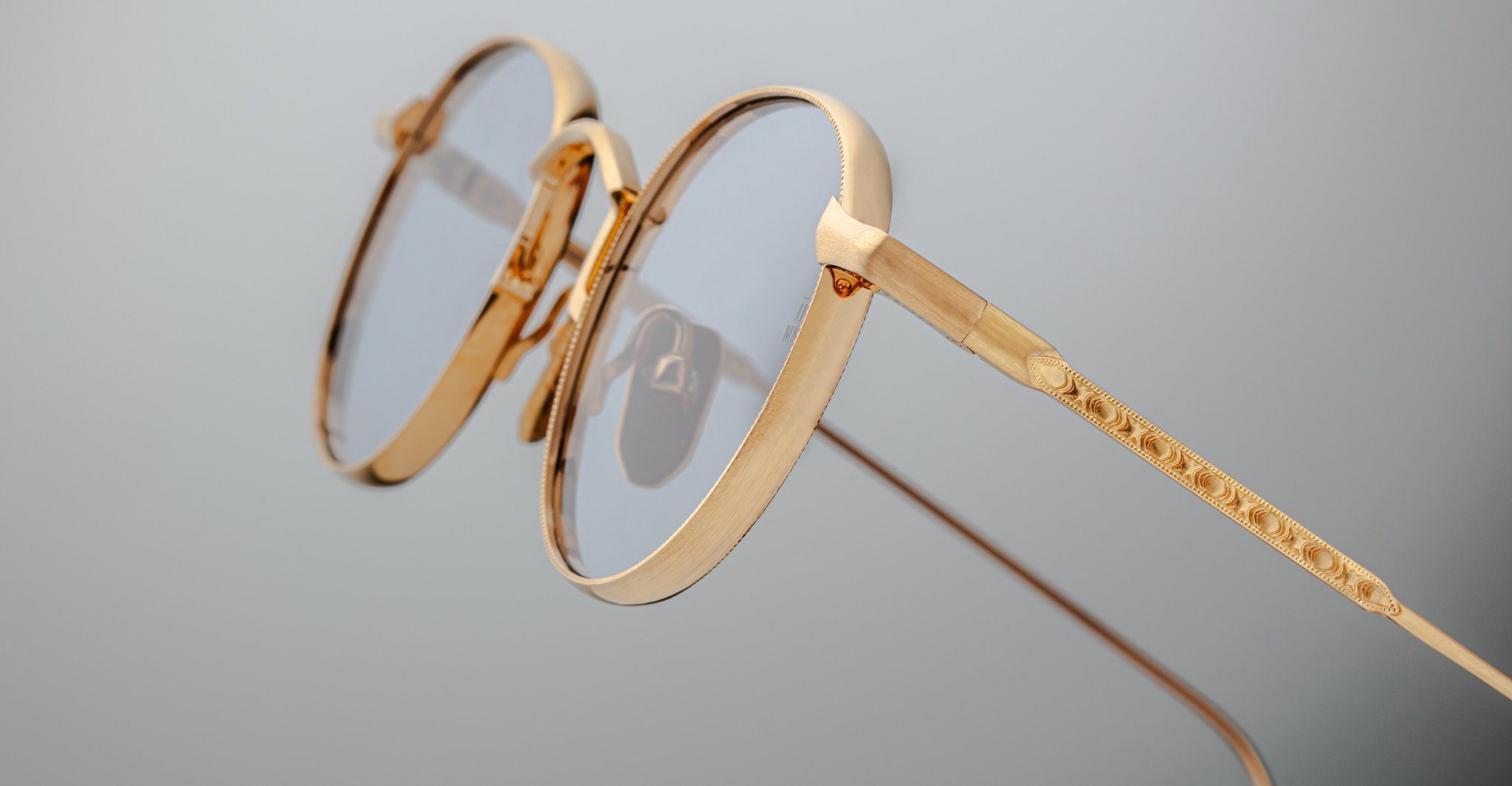 Close-up of Jacques Marie Mage Full Metal Jacket eyeglasses: gold-framed, round with clear lenses, featuring intricate detailing on the arms, set against a plain light gray background.