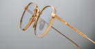 Close-up of Jacques Marie Mage Full Metal Jacket eyeglasses: gold-framed, round with clear lenses, featuring intricate detailing on the arms, set against a plain light gray background.