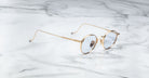 The Jacques Marie Mage Full Metal Jacket eyeglasses, featuring round gold frames with thin arms and clear lenses, rest on a white marble surface with grey veining.