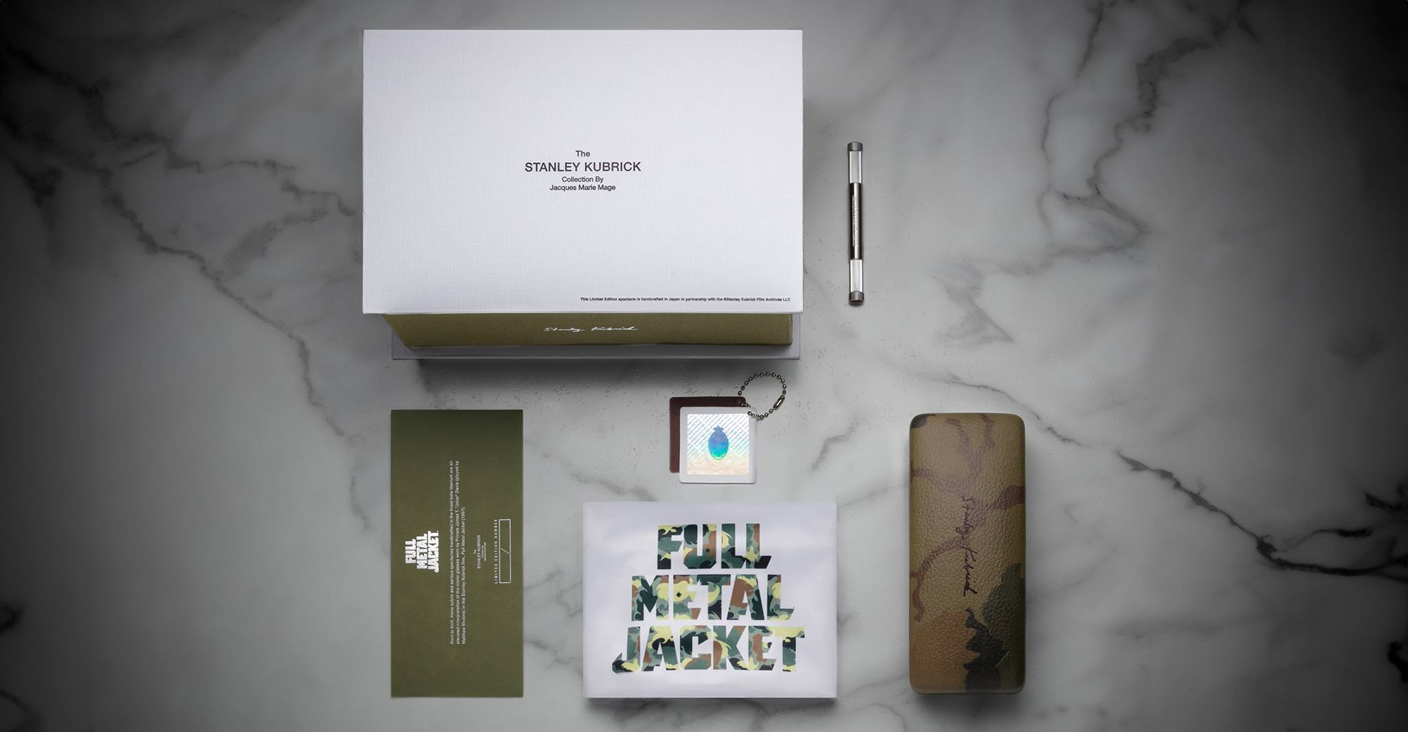 A flat lay of Jacques Marie Mage Full Metal Jacket merchandise on marble features a white box, metal screwdriver, camo glasses case, green envelope, keychain with blue drop, and a graphic card displaying the movie title.