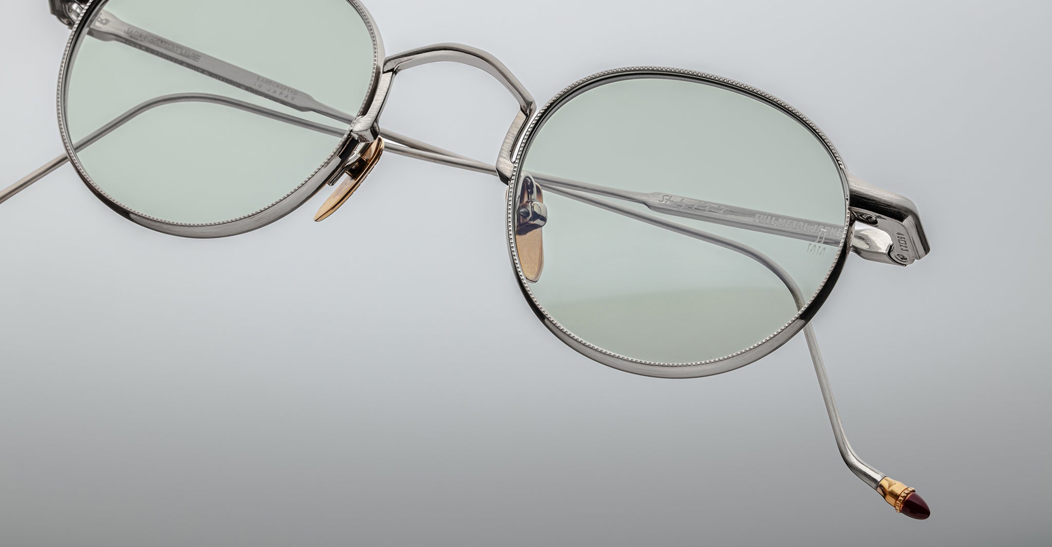 Close-up of Jacques Marie Mage Full Metal Jacket eyeglasses featuring stylish round silver frames, light green lenses, and gold and brown accents on the nose pads and temple tips, set against a smooth light gradient background.