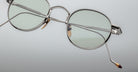 Close-up of Jacques Marie Mage Full Metal Jacket eyeglasses featuring stylish round silver frames, light green lenses, and gold and brown accents on the nose pads and temple tips, set against a smooth light gradient background.