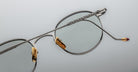 Close-up of Jacques Marie Mage Full Metal Jacket eyeglasses featuring silver metal round lenses, gold nose pads, a single extended temple arm with fine detailing, and a burgundy tip against a light gray background.