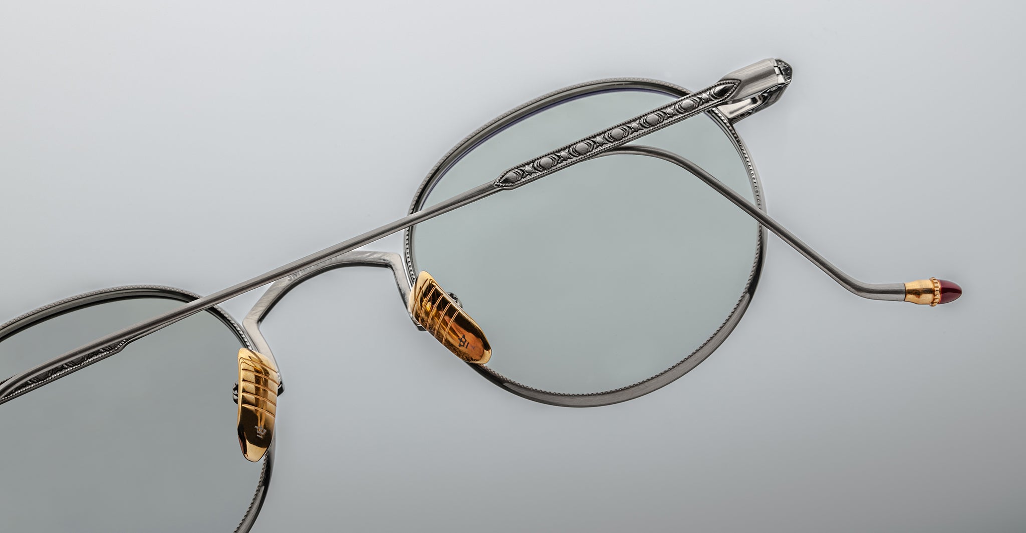 Close-up of Jacques Marie Mage Full Metal Jacket eyeglasses featuring silver metal round lenses, gold nose pads, a single extended temple arm with fine detailing, and a burgundy tip against a light gray background.