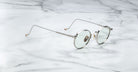 The Jacques Marie Mage Full Metal Jacket features round, silver metal frames with thin arms and clear lenses, displayed on a white marble surface.