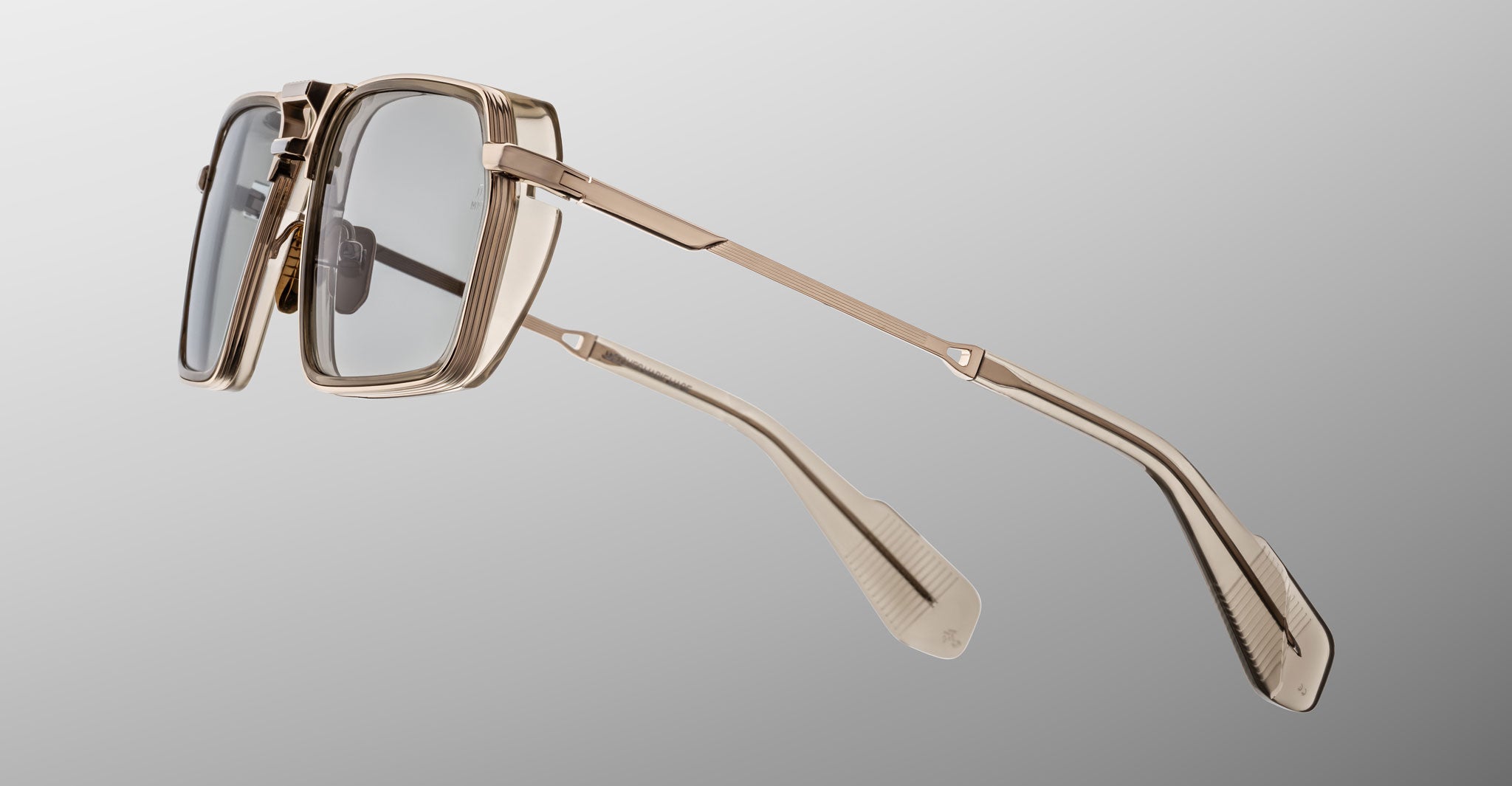 The Jacques Marie Mage GT-R eyeglasses feature a gold metal rectangular frame with clear lenses and detailed temple arms, displayed from a side angle against a light gray background.