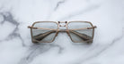 A pair of Jacques Marie Mage GT-R square, metal-framed eyeglasses with light-tinted lenses rests on a white marble surface.