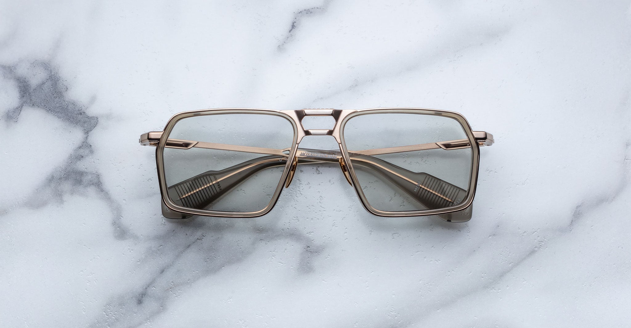 A pair of Jacques Marie Mage GT-R square, metal-framed eyeglasses with light-tinted lenses rests on a white marble surface.