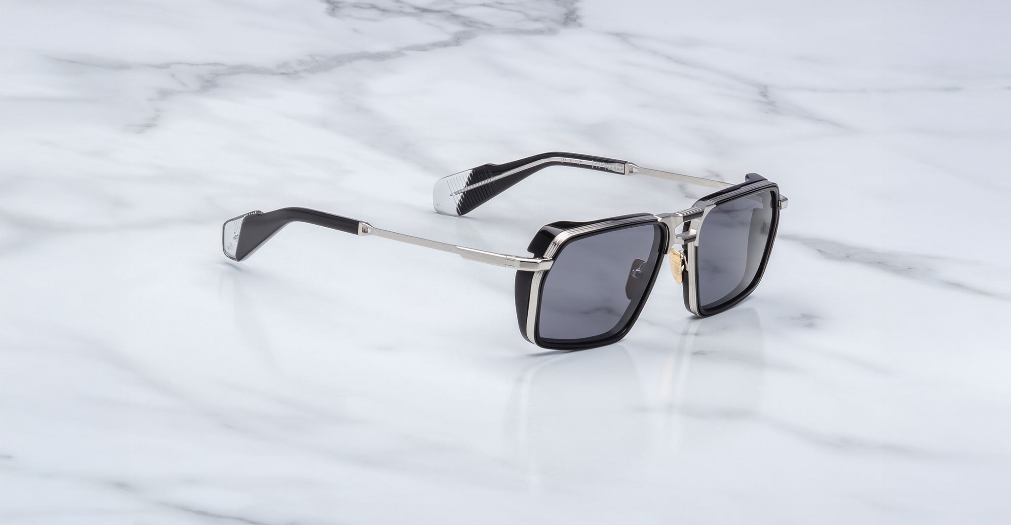 A pair of Jacques Marie Mage GT-R black square sunglasses with metal arms and dark lenses rests on a white marble surface.