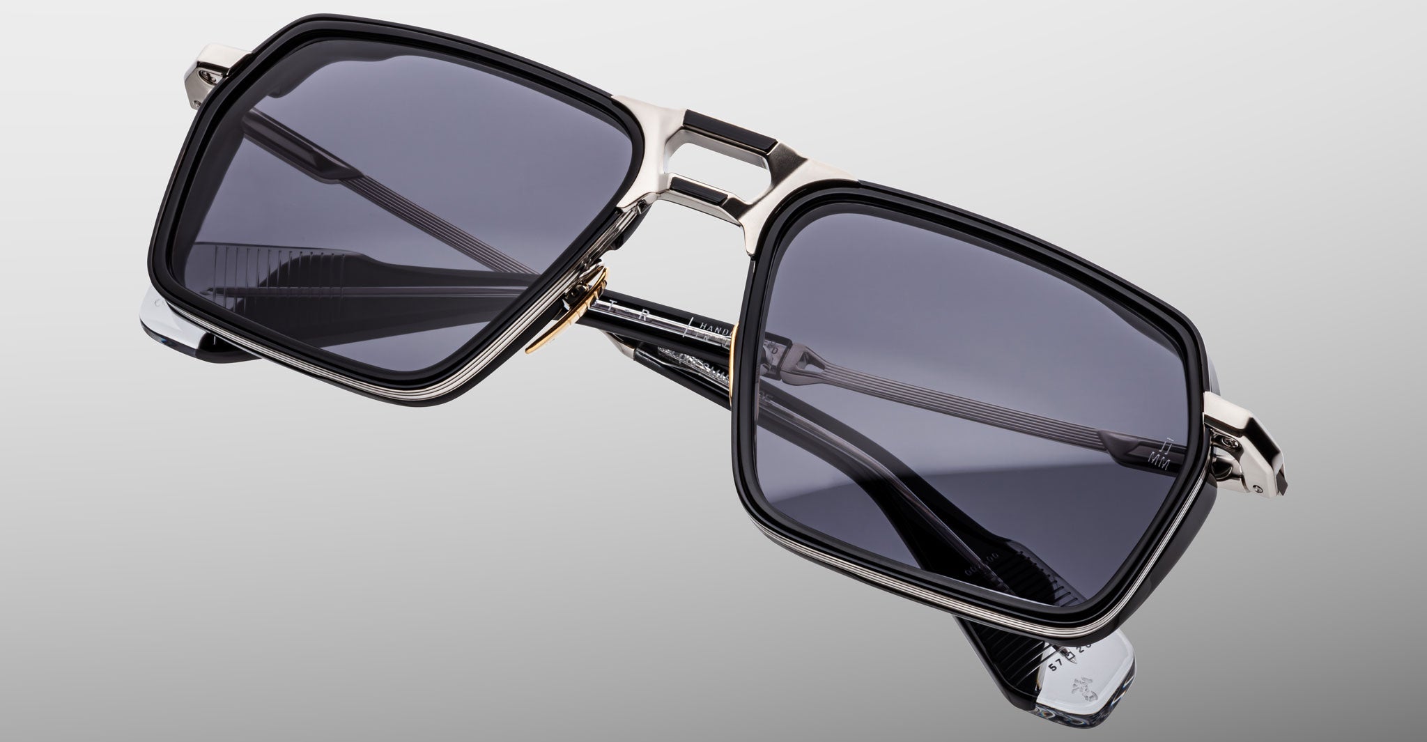 The Jacques Marie Mage GT-R features rectangular black frames, dark tinted lenses, and metallic accents on the bridge and temples, showcased against a neutral gradient background.