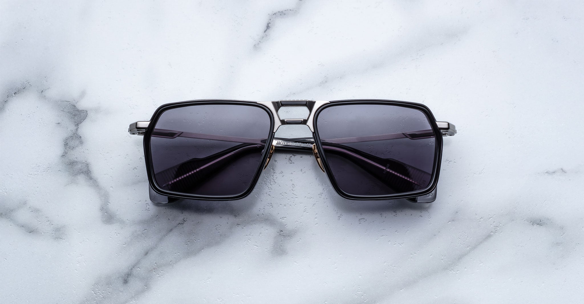 A pair of Jacques Marie Mage GT-R black square-framed sunglasses with dark lenses is displayed on a white marble surface.