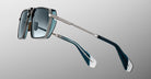 The Jacques Marie Mage GT-R sunglasses feature rectangular silver metal frames, blue-tinted lenses, and blue-and-silver arms, shown on a neutral gray background.