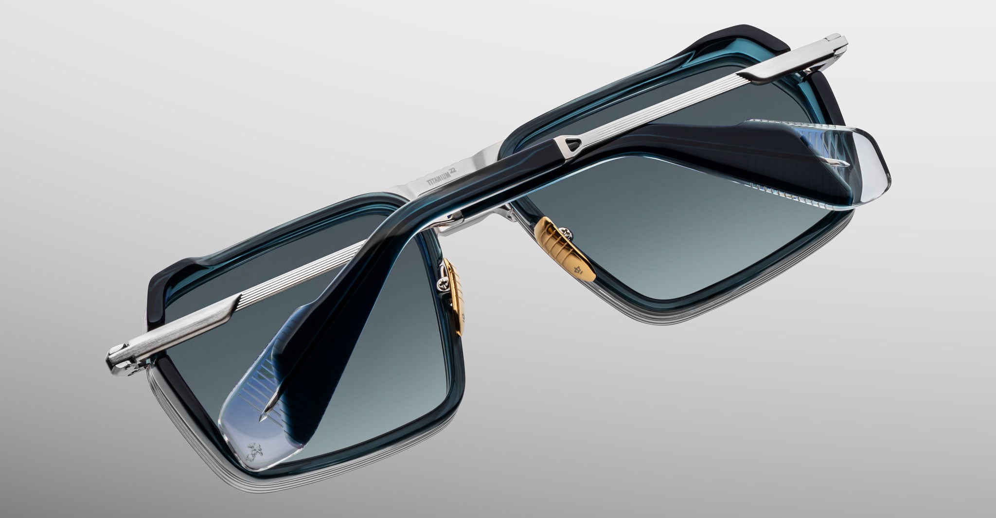 A close-up of Jacques Marie Mage GT-R sunglasses, featuring silver metal arms, transparent blue rectangular frames, and tinted lenses, displayed from an angled overhead view on a light gray background.