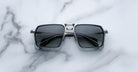 A pair of Jacques Marie Mage GT-R square, metal-framed sunglasses with dark lenses rests on a white marble surface.