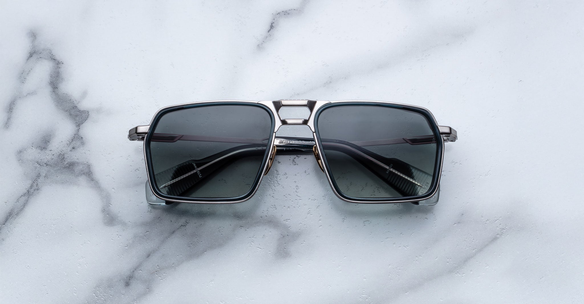 A pair of Jacques Marie Mage GT-R square, metal-framed sunglasses with dark lenses rests on a white marble surface.