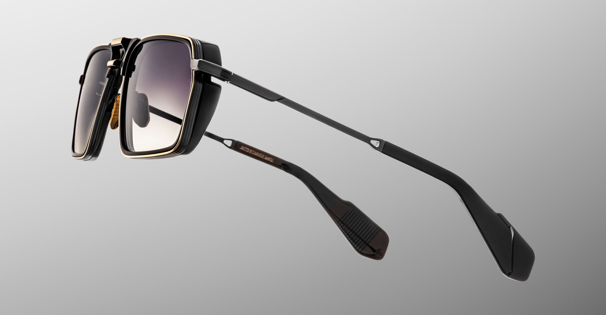 A close-up of Jacques Marie Mage GT-R black sunglasses with gold accents and rectangular lenses, set against a plain gray background. Sleek, angled temples highlight the distinctive hinge and earpiece design.