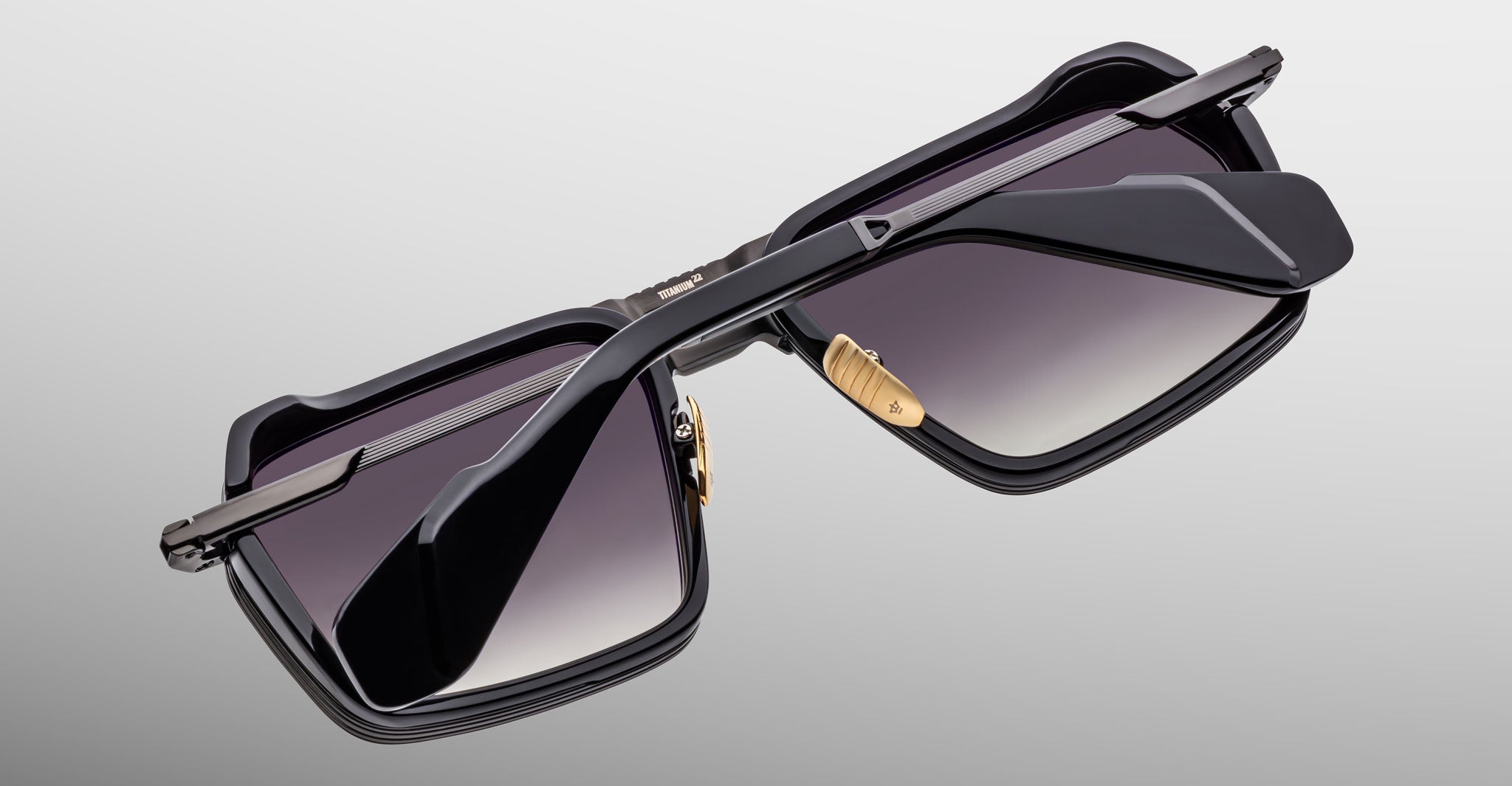 A close-up of the Jacques Marie Mage GT-R sunglasses shows rectangular gradient lenses, gold nose pads, and black frames set upside down on a light grey background, emphasizing their distinctive frame and side arms.