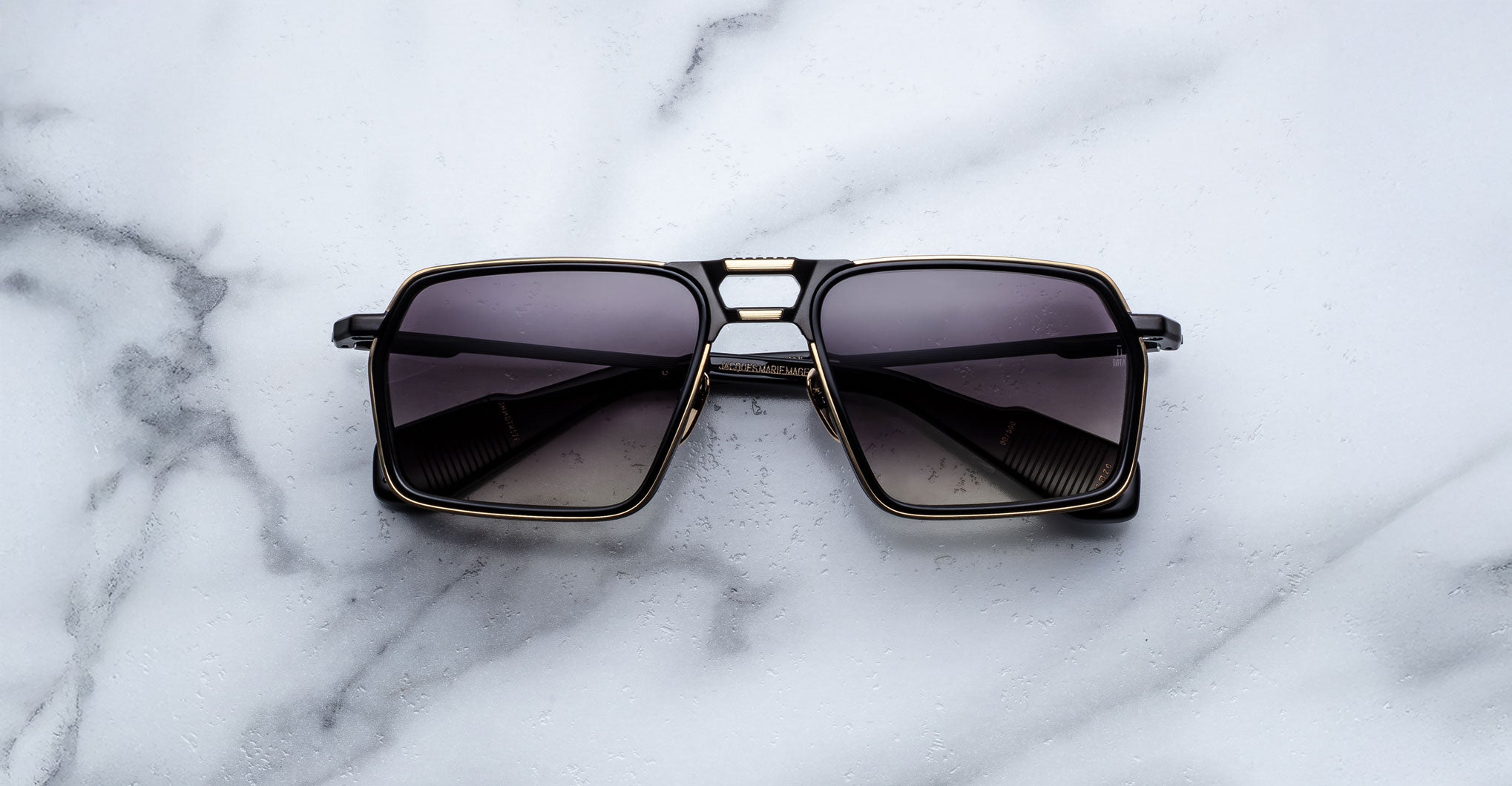 A pair of Jacques Marie Mage GT-R black square-framed sunglasses with dark tinted lenses rests on a white marble surface.