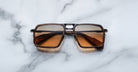 A pair of Jacques Marie Mage GT-R rectangular brown sunglasses with dark lenses rests on a white marble surface.