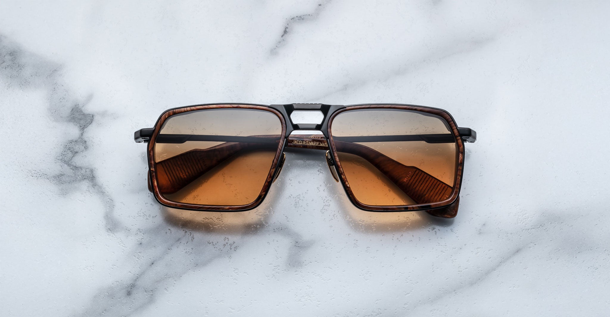 A pair of Jacques Marie Mage GT-R rectangular brown sunglasses with dark lenses rests on a white marble surface.