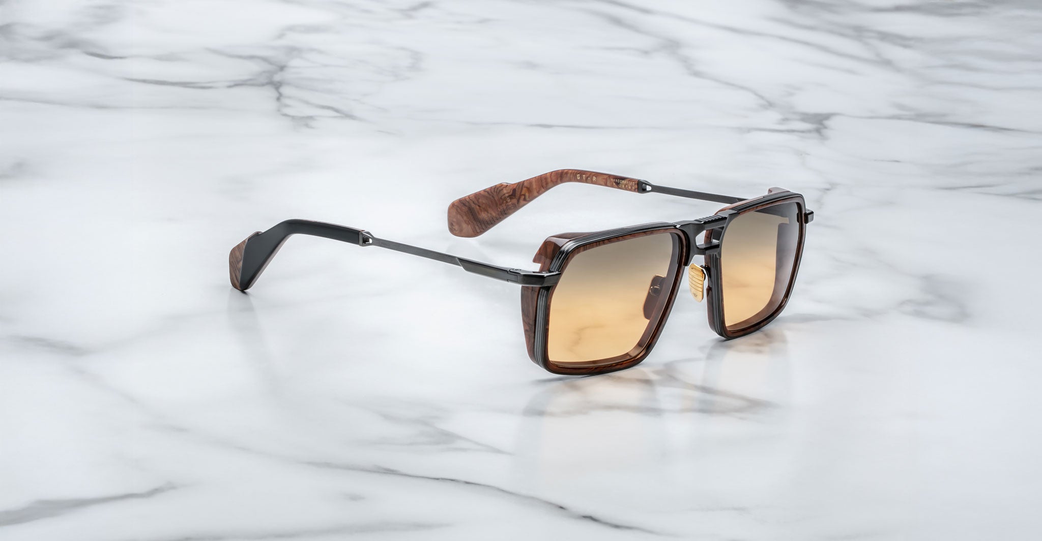 A pair of Jacques Marie Mage GT-R sunglasses featuring brown square frames, gradient amber lenses, and wood-textured arms, displayed on a white marble surface.