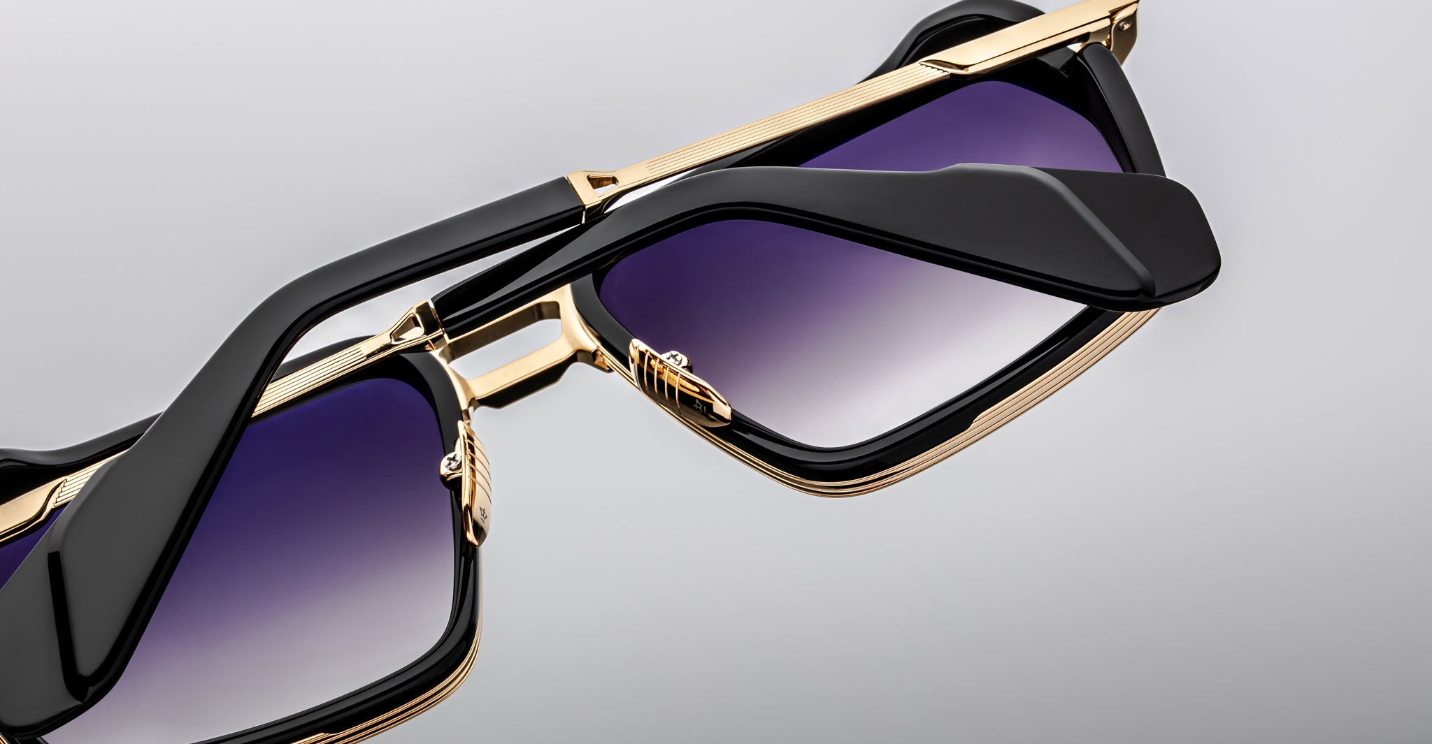 Close-up of two Jacques Marie Mage GT-R sunglasses with gold frames, black arms, and gradient purple lenses, one stacked atop the other against a plain grey background.