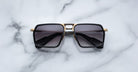 Jacques Marie Mage GT-R rectangular sunglasses with dark lenses, black frames, and gold accents rest on a white marble surface.