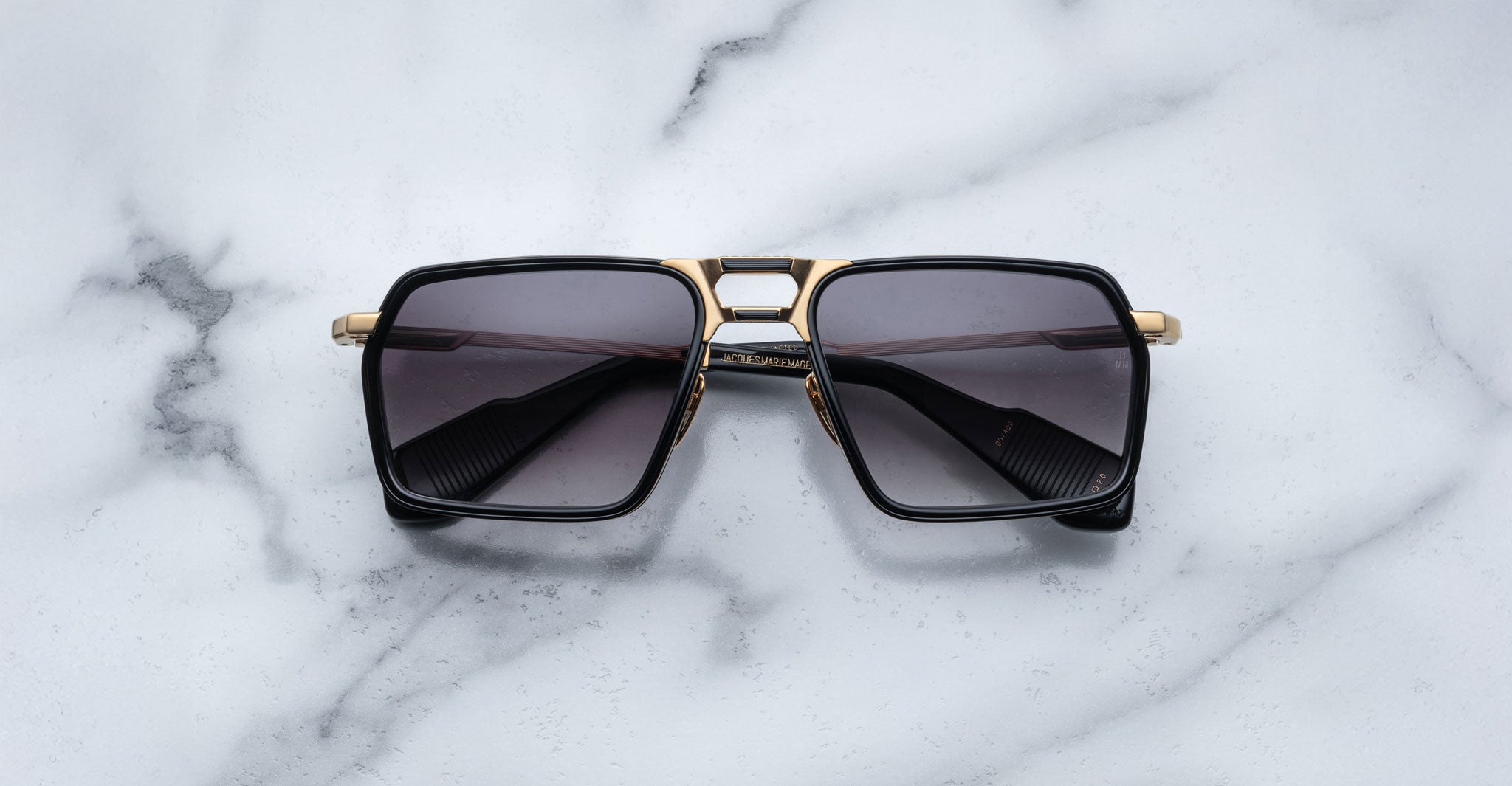 Jacques Marie Mage GT-R rectangular sunglasses with dark lenses, black frames, and gold accents rest on a white marble surface.
