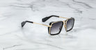 The Jacques Marie Mage GT-R sunglasses feature rectangular black frames with gold accents, dark lenses, and subtly textured temples near the ear tips, shown resting on a white marble surface.