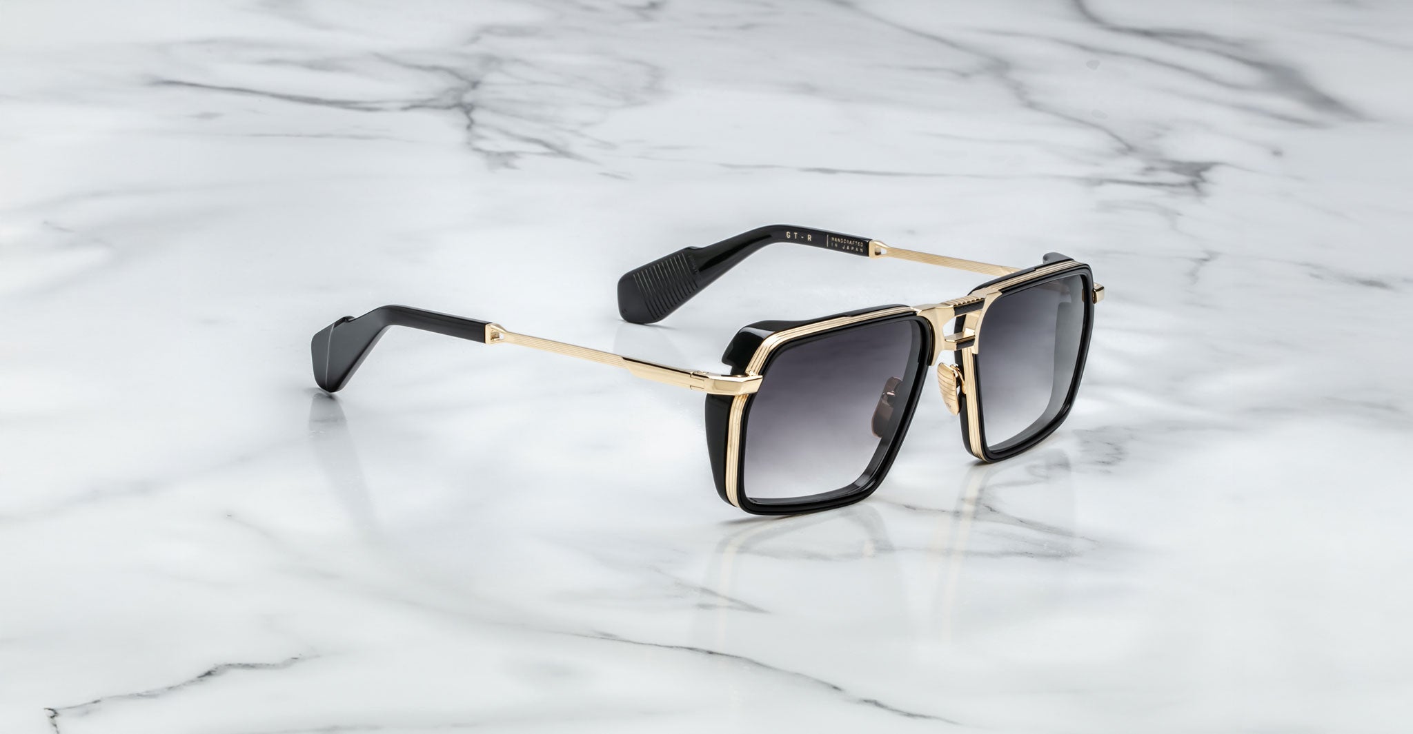 The Jacques Marie Mage GT-R sunglasses feature rectangular black frames with gold accents, dark lenses, and subtly textured temples near the ear tips, shown resting on a white marble surface.