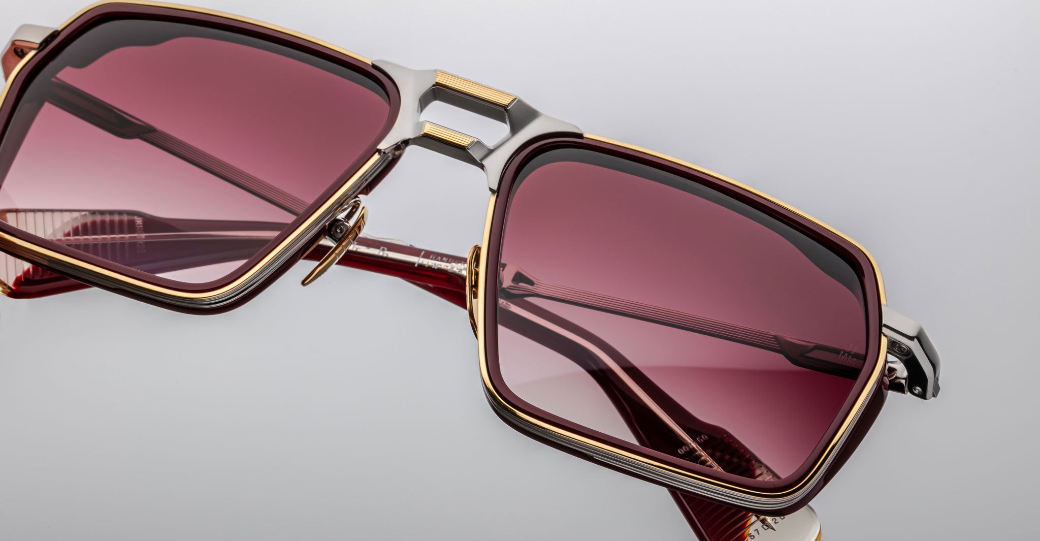 Close-up of Jacques Marie Mage GT-R sunglasses featuring square frames, gold and silver detailing, pink tinted lenses, and metallic-accented temples set against a plain light gray background.
