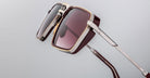 Close-up of Jacques Marie Mage GT-R sunglasses: square burgundy frames with gold and silver accents on the frame and temples, displayed against a plain gray background.