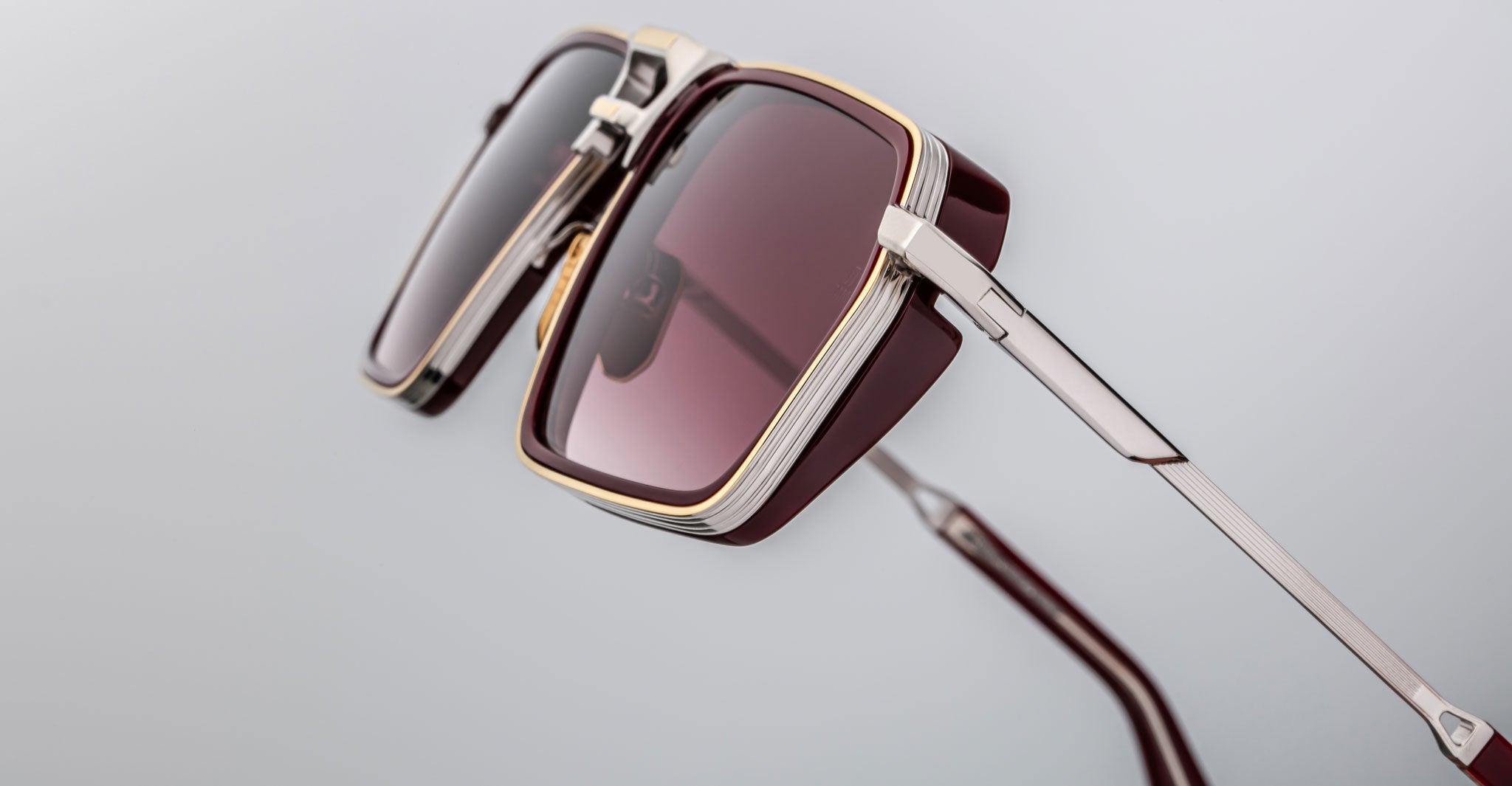 Close-up of Jacques Marie Mage GT-R sunglasses: square burgundy frames with gold and silver accents on the frame and temples, displayed against a plain gray background.