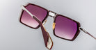 A close-up of Jacques Marie Mage GT-R sunglasses with gradient purple lenses, metallic gold nose pads, and a thick dark red arm, captured at an angle against a plain gray background.